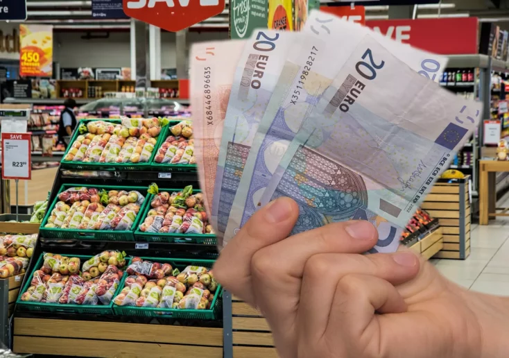 Symbolfoto zu einem Beitrag von 5min.at: Eine Hand hält Bargeld vor einem Gemüseregal im Supermarkt