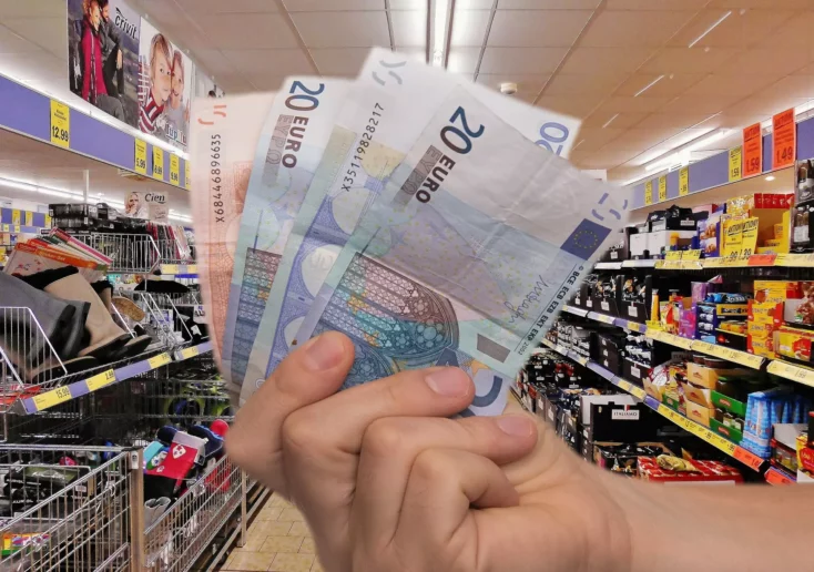 Symbolfoto zu einem Beitrag von 5min.at: Eine Hand hält Geldscheine, im Hintergrund Supermarktregale