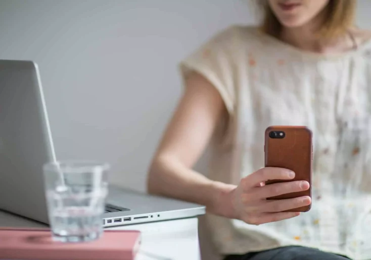 Symbolfoto zu einem Beitrag von 5min.at: Eine Frau sitzt an einem Tisch und schaut auf ihr Handy, auf dem Tisch steht ihr Laptop