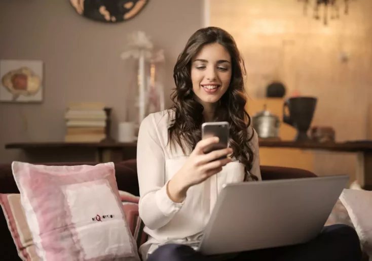 Symbolfoto zu einem Beitrag von 5min.at: Eine lächelnde Frau sitzt auf einem Sofa und schaut auf ihr Handy, auf ihrem Schoß ist ein Laptop