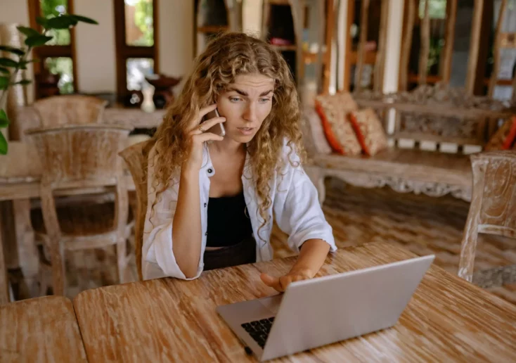 Symbolfoto zu einem Beitrag von 5min.at: Eine Frau sitzt an einem Tisch, auf dem ein Laptop steht, und telefoniert