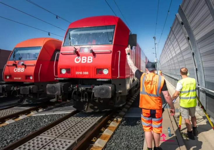 Symbolfoto von 5min.at: Bauarbeiter stehen zwischen einem Zug und einer Schallschutzwand.