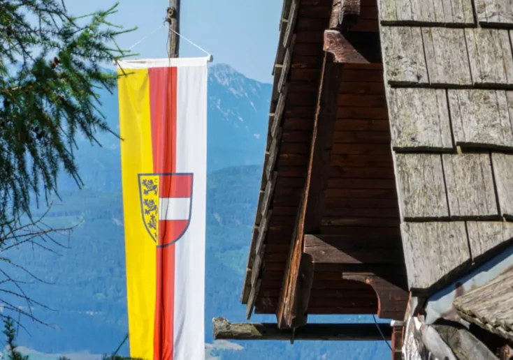 Ein Bild auf 5min.at zeigt eine Kärntnerflagge vor der wunderschönen Berglandschaft Kärntens. Rechts steht eine Hütte.
