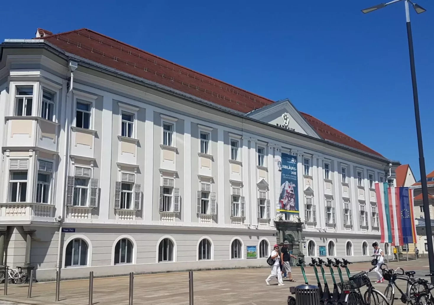Ein Bild auf 5min.at zeigt die Außenfassade des Klagenfurter Rathauses, davor schlendern Spaziergänger durch die Stadt. Der Himmel ist blau.