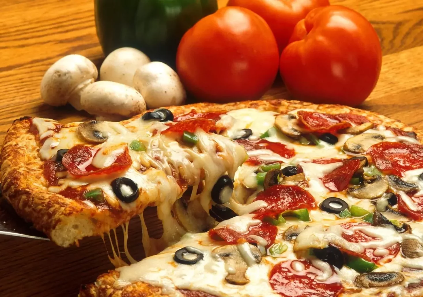 Ein Bild auf 5mib.at zeigt eine Pizza. Dahinter liegen Champignons und Tomaten. Ein Stück der Pizza wird gerade angehoben.