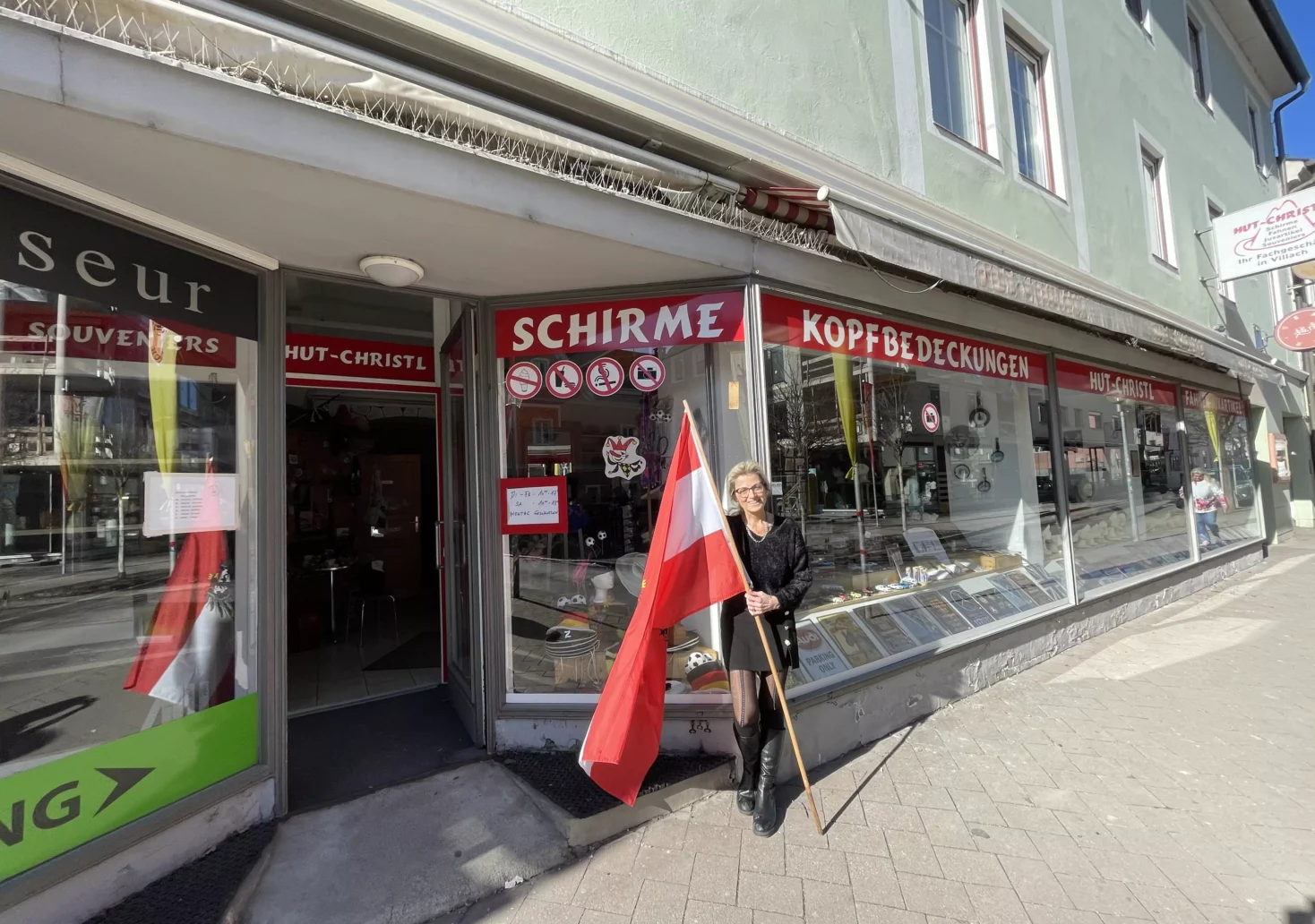 Bild auf 5min.at zeigt ein Hutgeschäft in Villach von außen, davor eine Frau mit Österreich-Flagge.