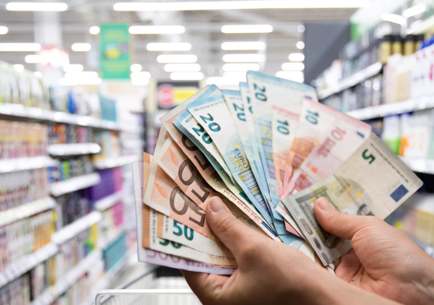 Ein Bild auf 5min.at zeigt einen Bündel Geldscheine vor einem Supermarktregal.