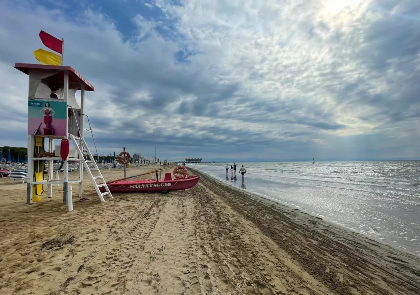 Ein Bild auf 5min.at zeigt Touristen, die am Strand in Lignano spazieren gehen. Rechts rauschen Wellen vom Meer heran.