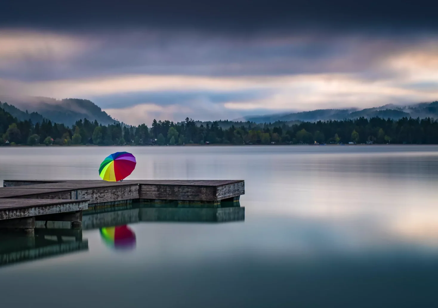 Ein Bild auf 5min.at zeigt dunkle Wolken über dem Faaker See in Kärnten. Auf einem Steg steht ein aufgespannter Regenbogenschirm.
