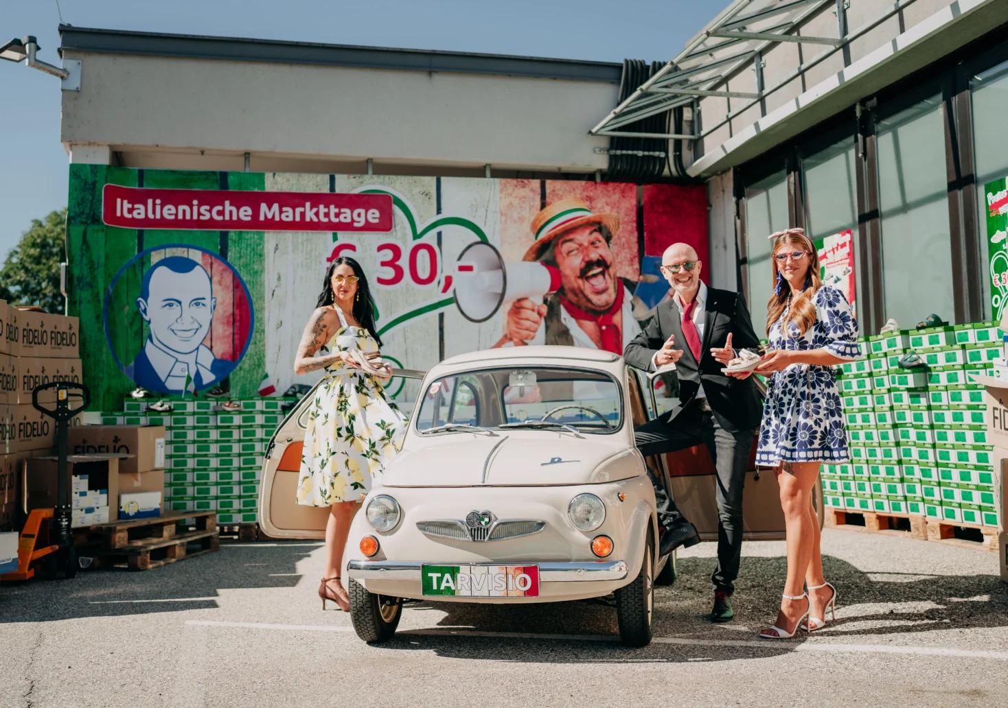 Drei Personen stehen neben einem Oldtimer vor einem Ranacher-Standort. Im Hintergrund wirbt ein großes Plakat für die „Italienischen Markttage“ und Pantoffel um 30 Euro.